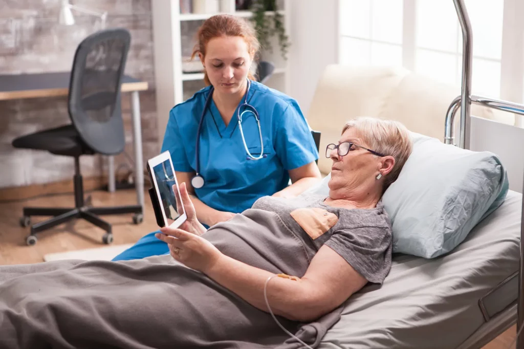 Old woman in nursing home sitting in bed using tablet with female nurse sitting next to bed. Our Kansas City personal injury lawyers fight for those abused or neglected in nursing homes. 